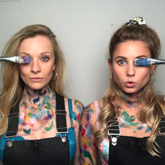 two women covered in paint, holding paint brushes to their eyes