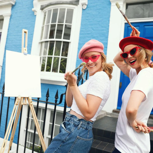 two women holding wearing berets, holding paint brushes in front of a blank canvas 