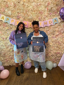 two women hold seascape paintings at paint and sip birthday event